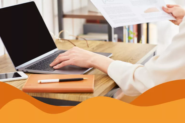 image of a woman wearing a white long sleeve on her laptop with a dark screen, also holding paperwork and with a book at the desk in an article about how strong it support improves employee productivity